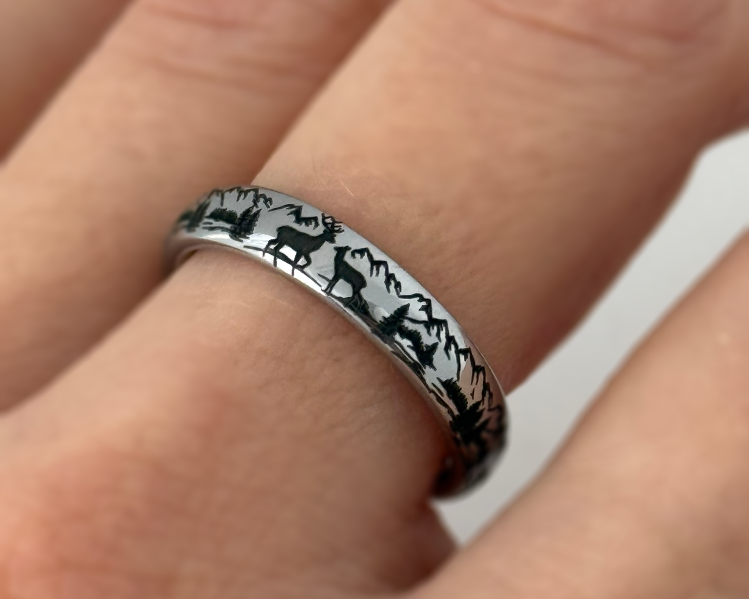 Nature inspired tungsten ring with detailed laser engraving of mountains and deer, worn on a finger.