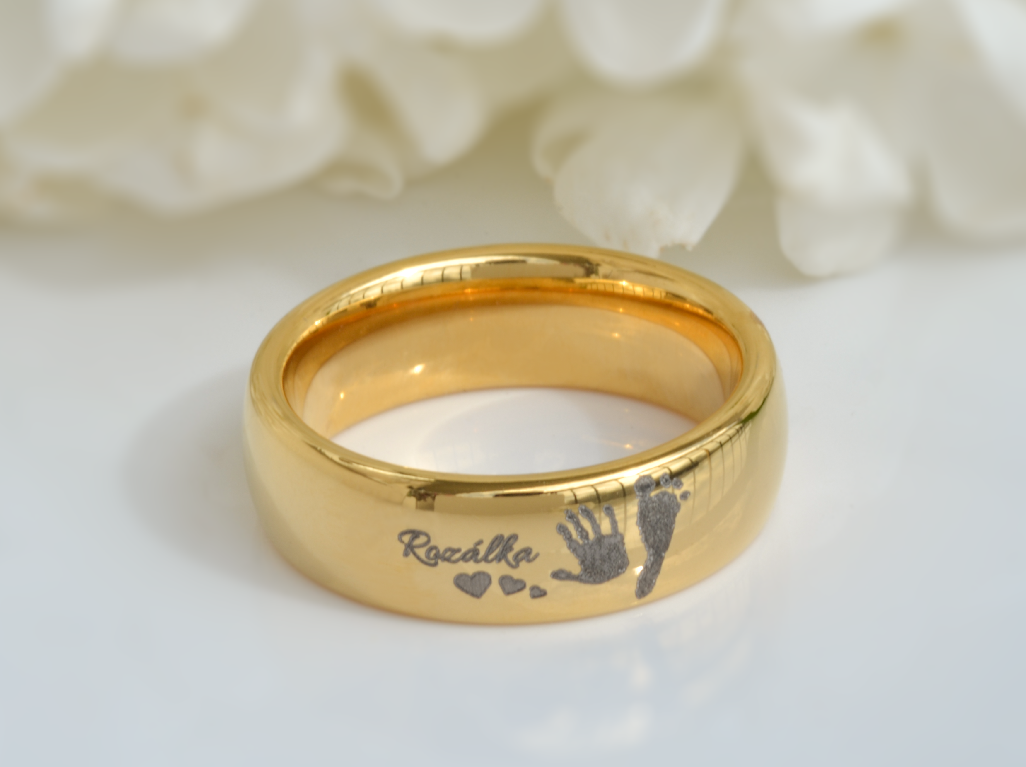  A golden tungsten ring named 'Eternal Imprint' displayed against a soft, white floral background. The ring features a personalized engraving of a child's handprint and footprint alongside small hearts, capturing a sentimental and enduring memory in a durable piece of jewelry.