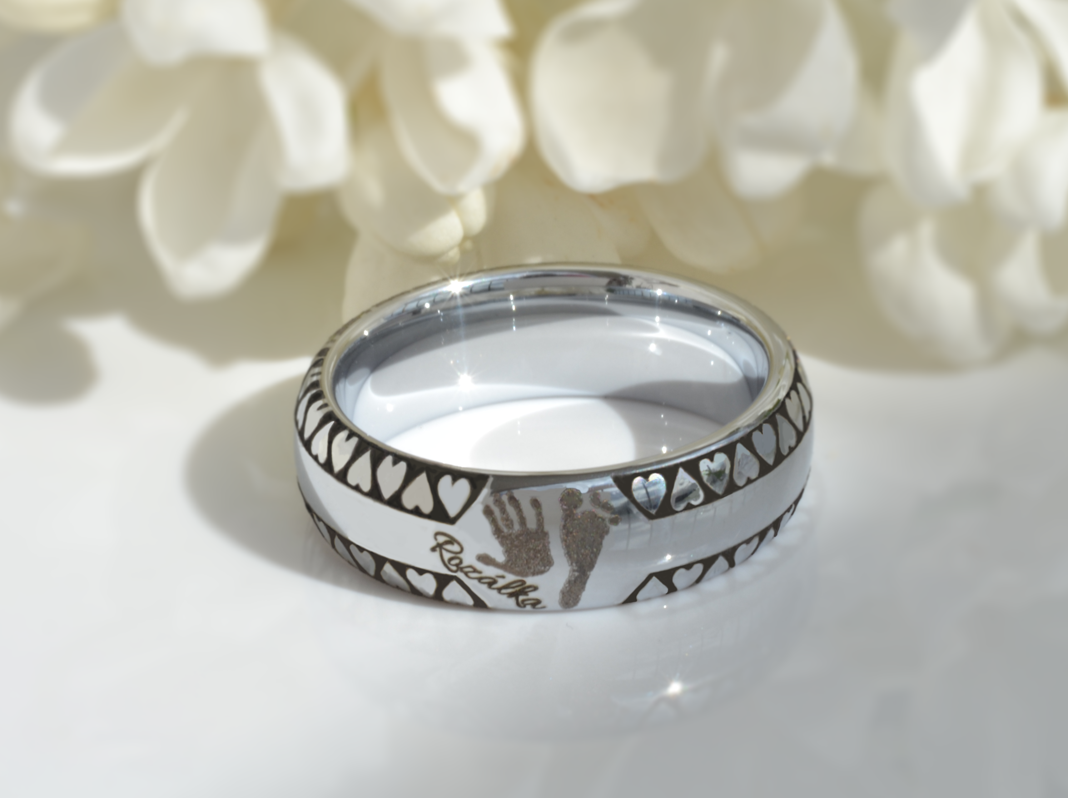 A white gold tungsten ring, elegantly displayed among white flowers, is adorned with a continuous heart pattern and a personalized engraving of a child's handprint. This ring combines affectionate symbols with the name 'Rozálka' etched alongside the heart and hand motifs, representing a meaningful gesture of love and remembrance.