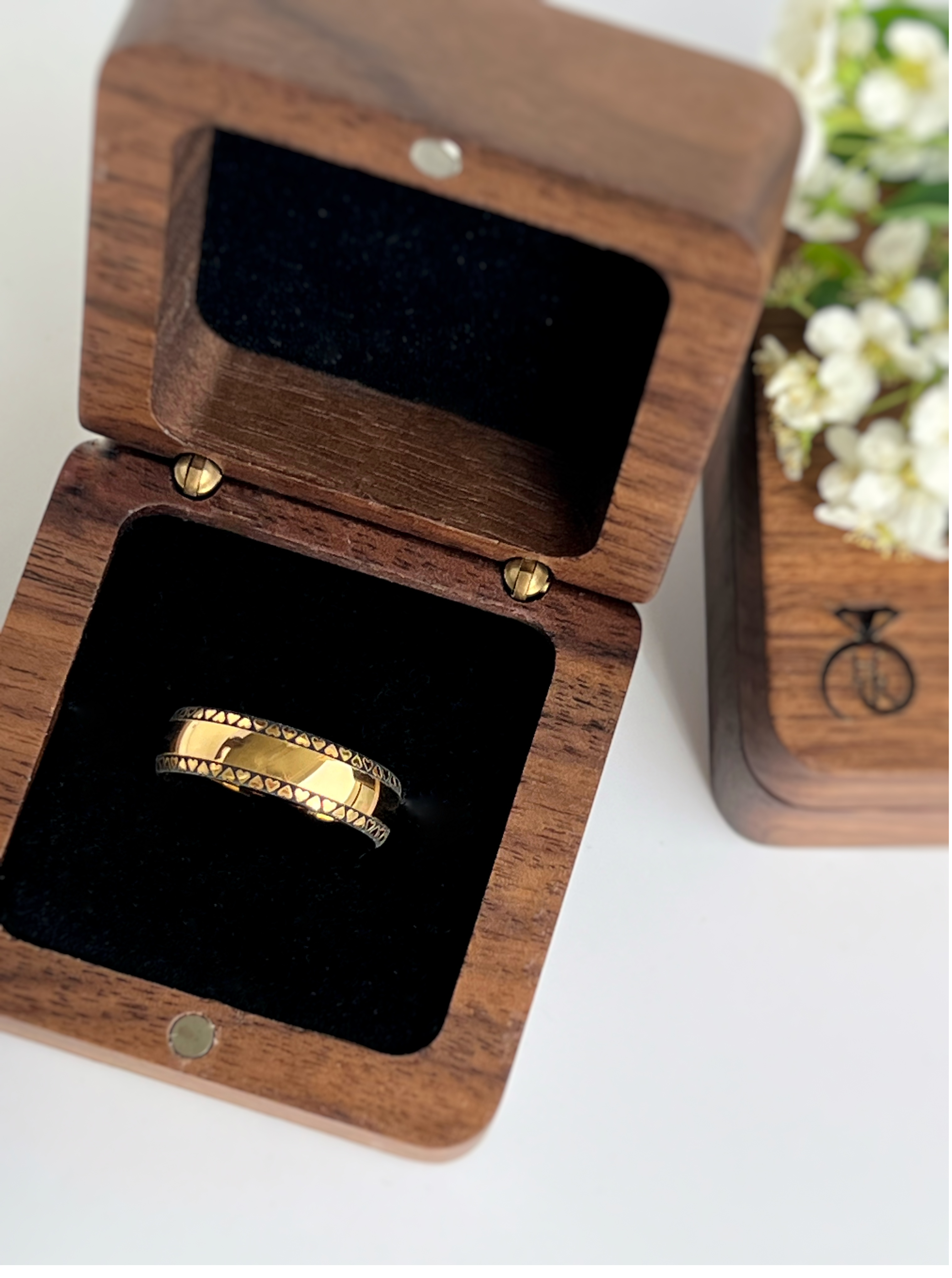 Gold 'Eternal Love Band' with a pattern of black hearts displayed in a wooden ring box with a reflective black interior, positioned next to white flowers, offering an elegant presentation.