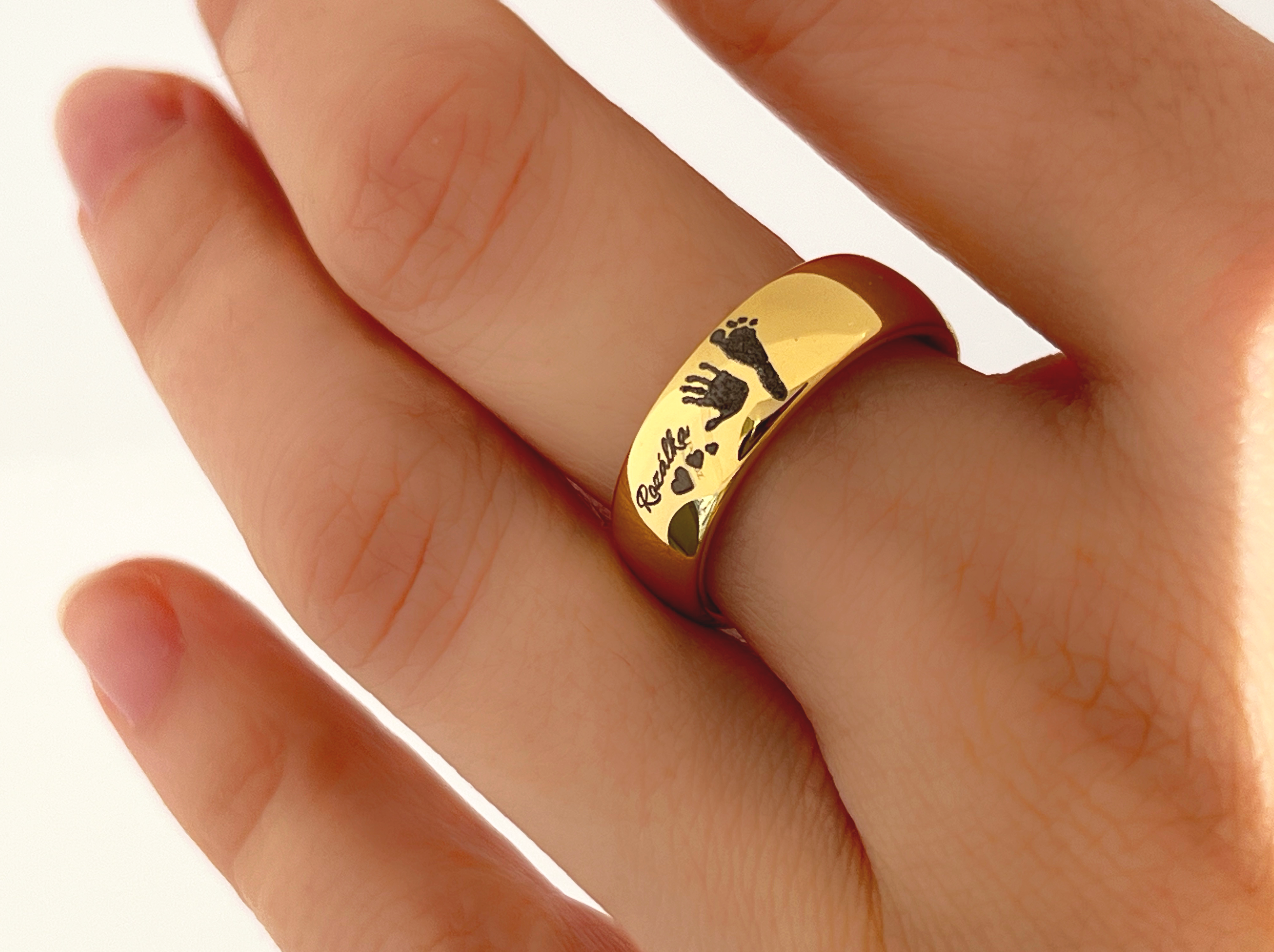 A hand wearing a gold tungsten ring named Eternal Imprint, which is engraved with a genuine silver-grey handprint and footprint of a child. The personalized engravings on the ring make it a meaningful piece, symbolizing the lasting connection between a parent and their child.