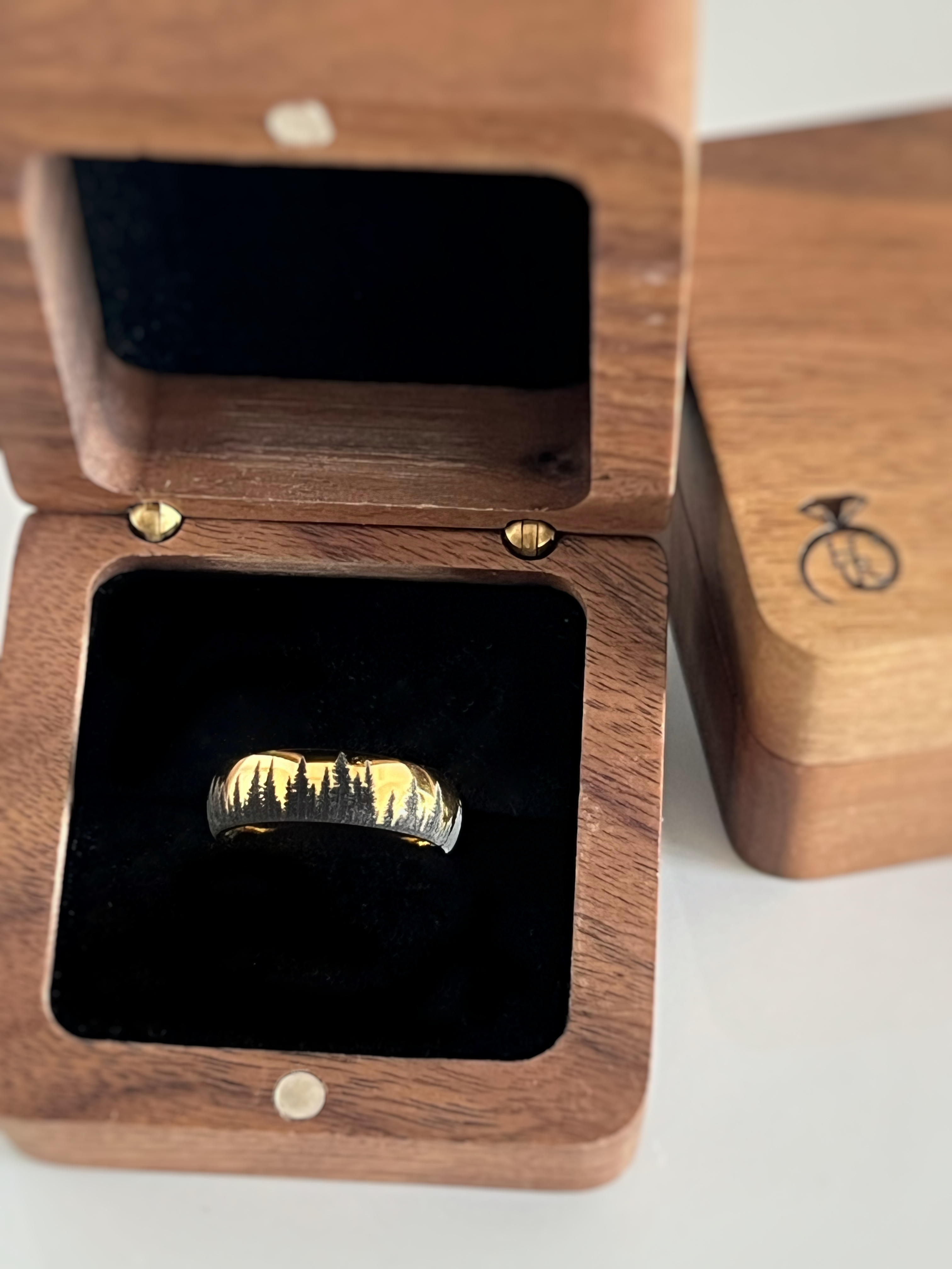 Gold tungsten ring with an engraved silhouette of an evergreen forest, displayed in an open wooden ring box with a black interior.