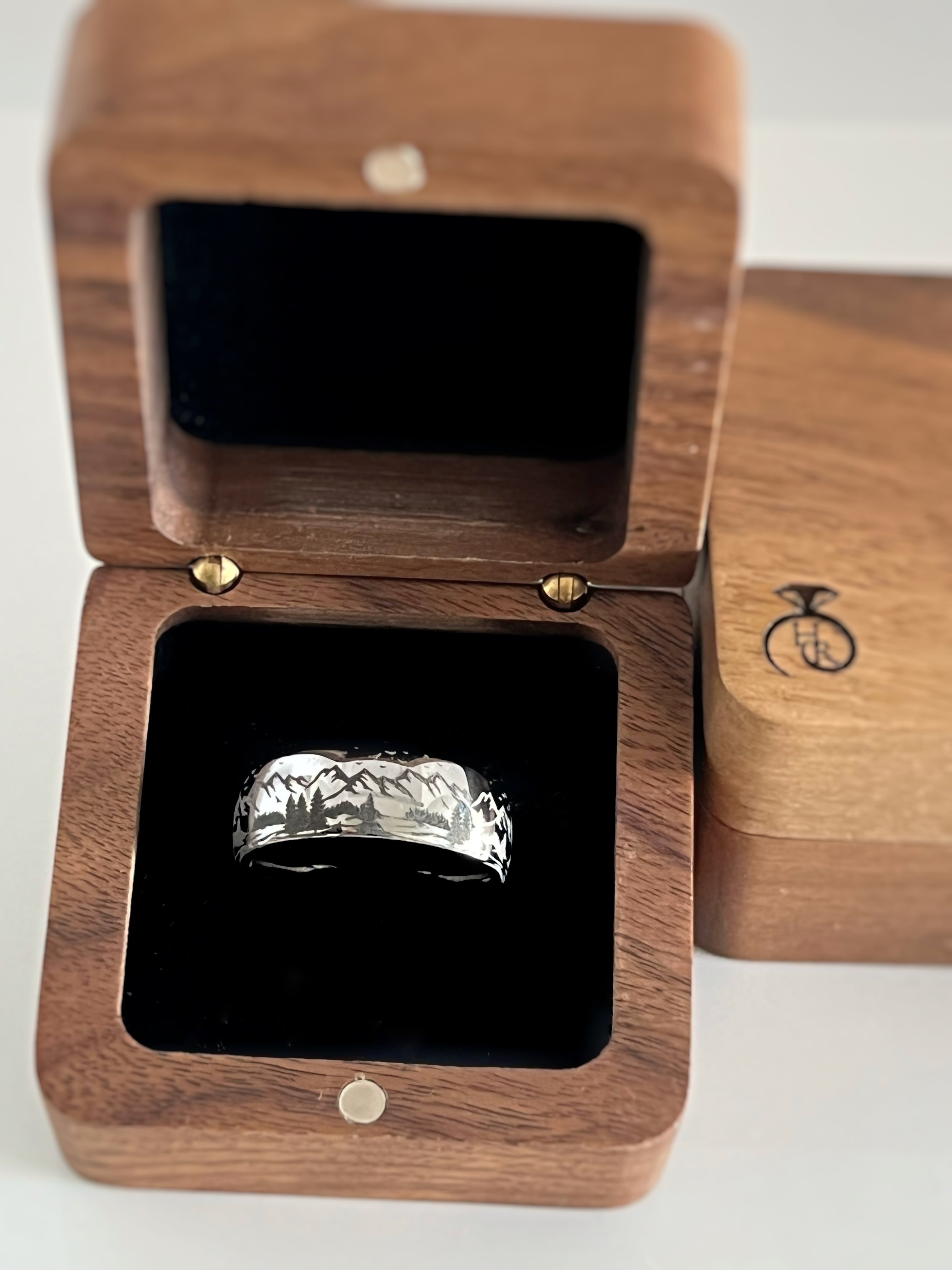 Silver tungsten ring with an engraved mountain range design, displayed in an open wooden ring box with a black interior.