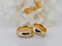 Pair of gold tungsten rings with mountain design symbolizing love and adventure, styled with flowers.