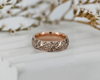 Elegant rose gold tungsten ring with engraved oak leaf pattern placed on a marble surface with dried flowers. Nature inspired wedding band with warm romantic aesthetic.
