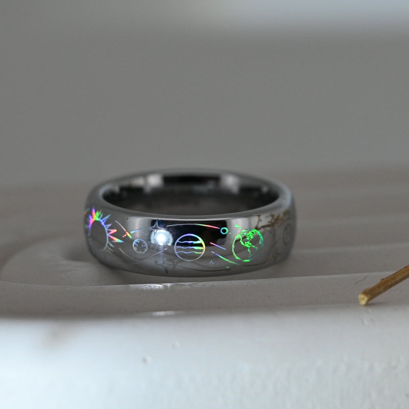 Silver-tone Celestial Orbit tungsten ring with holographic planets, stars and orbital lines, resting on a pale display surface.