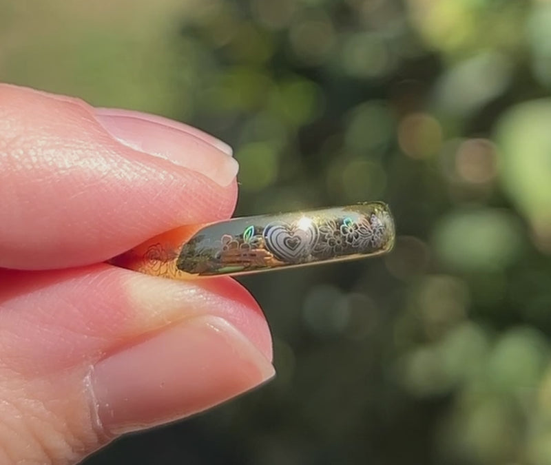 Close-up of a holographic tungsten ring held between fingers in sunlight, revealing a heart and intricate floral design with shifting rainbow colors on a polished gold band.