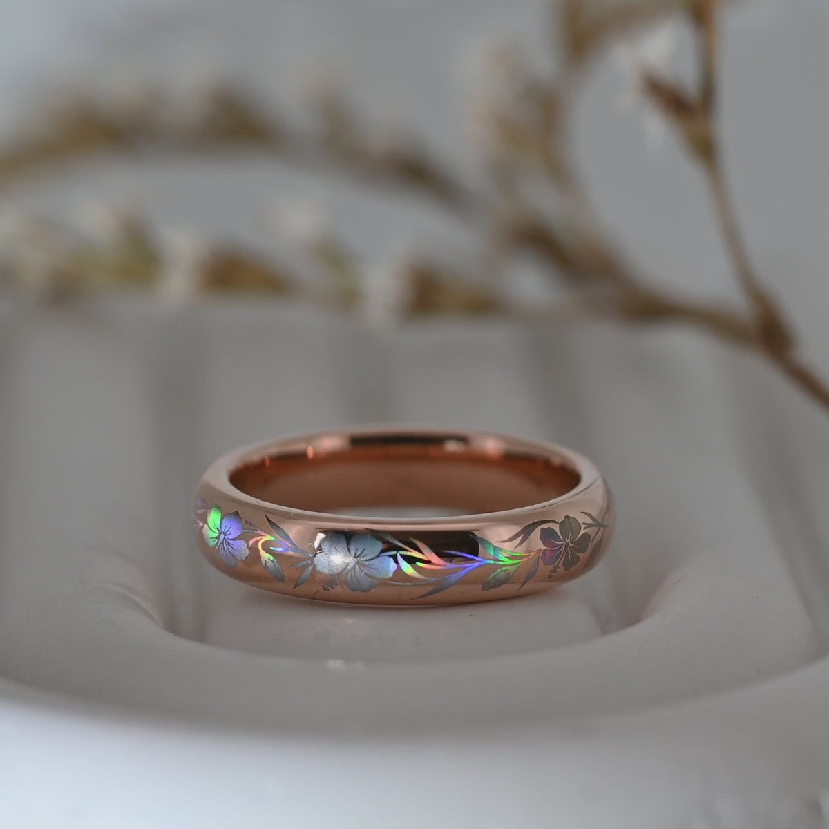Video of a rose-gold Hibiscus Glow tungsten ring on a pale display surface, showing holographic hibiscus flowers and flowing leaves.