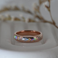 Video of a rose-gold Hibiscus Glow tungsten ring on a pale display surface, showing holographic hibiscus flowers and flowing leaves.