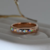 Rose-gold Sakura Glow tungsten ring with holographic sakura blossoms and flowing leaves, resting on a pale display surface.