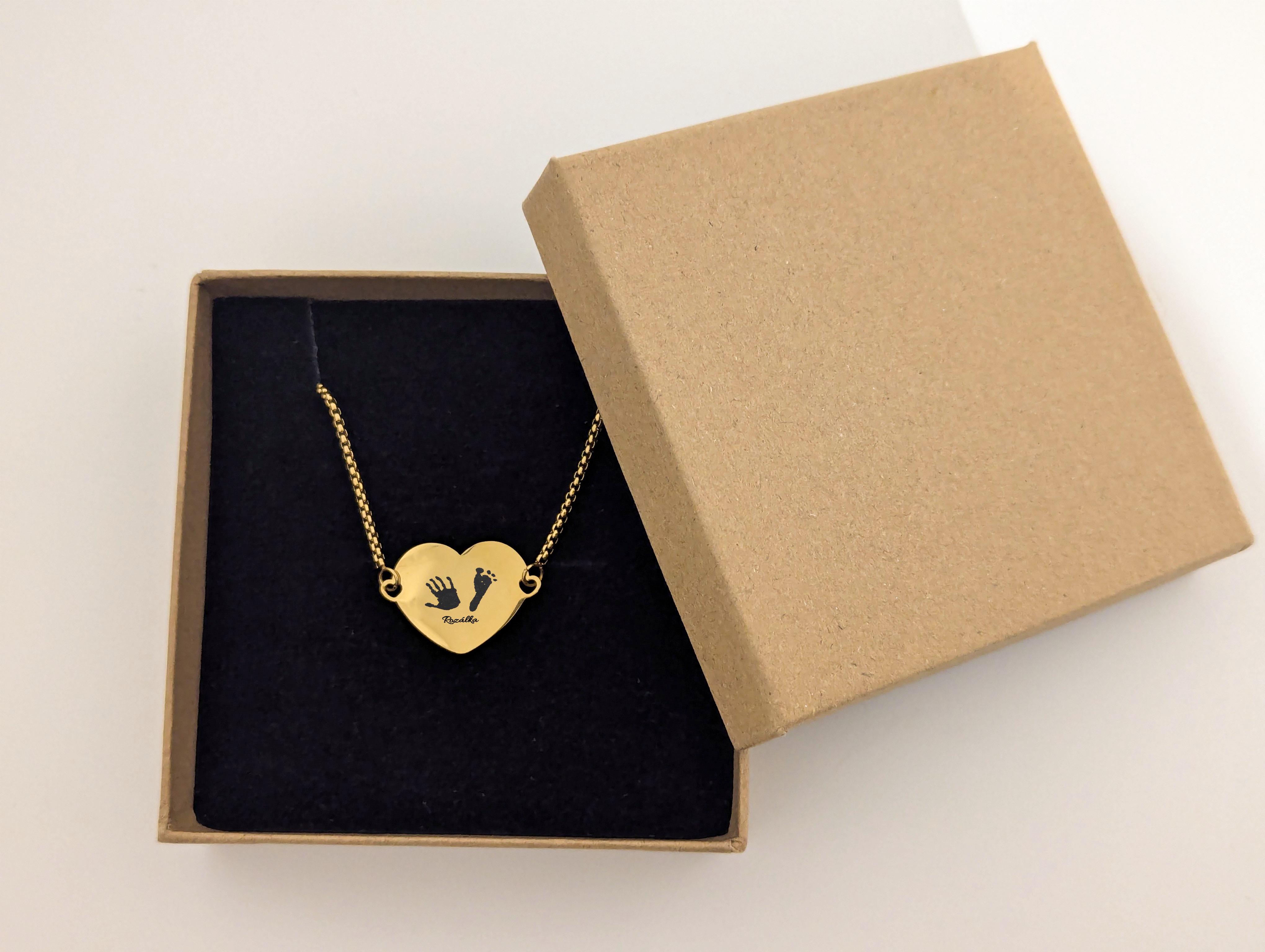 A gold-plated stainless steel heart-shaped bracelet displayed in a simple brown gift box with a black velvet lining. The bracelet features small engraved designs of a handprint and a footprint alongside the engraved name "Rosalie." The durable yet elegant gold-plated chain complements the personalized pendant, making it a sentimental and long-lasting keepsake.